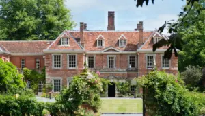 A stately red brick mansion with a tiled roof surrounded by lush green gardens.