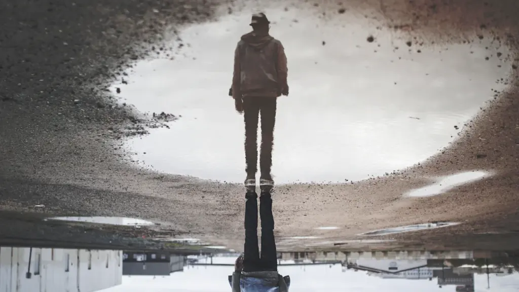 A person stands beside a puddle reflecting their silhouette in an inverted perspective.
