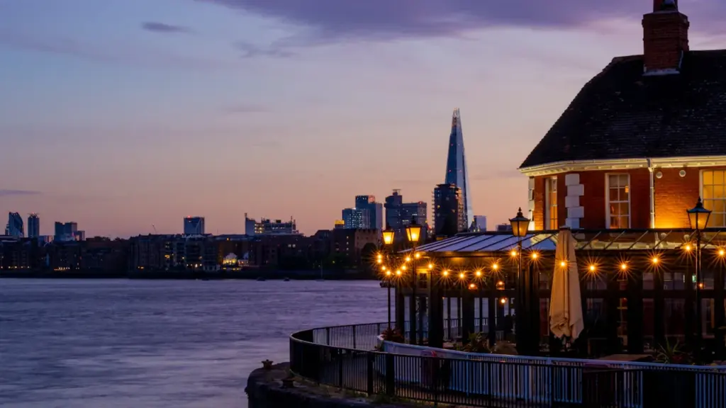 Illuminated riverside pub with warm lights against the London city skyline at twilight.