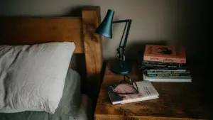 Books and reading glasses on a bedside table with a lamp.
